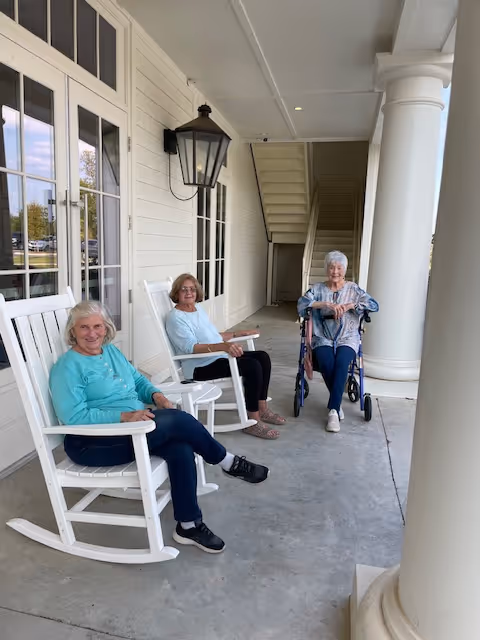 Three elderly women sitting on a covered porch of a building. Two women are seated in white rocking chairs, and one woman is sitting in a wheelchair. The porch has large white columns and a wall-mounted lantern light fixture. There are windows and a door along the wall behind them.