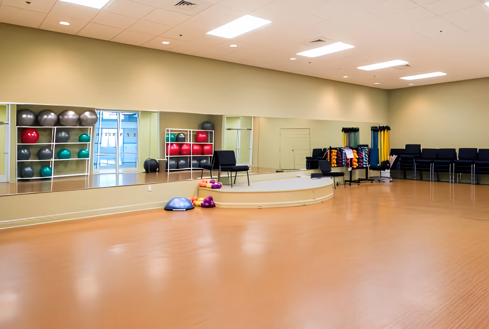 A spacious exercise or activity room with a wooden floor, large wall mirrors, and shelves holding various exercise balls and equipment. There are chairs arranged along the wall and a small raised platform with more exercise equipment nearby.