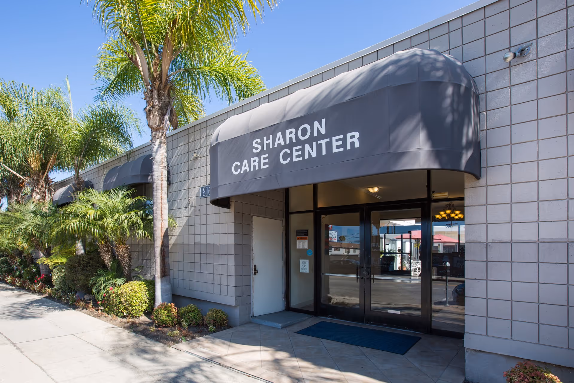 Exterior view of Sharon Care Center building entrance with a gray awning displaying the facility name. The building has a tiled facade and is surrounded by palm trees and shrubs along the sidewalk under a clear blue sky.