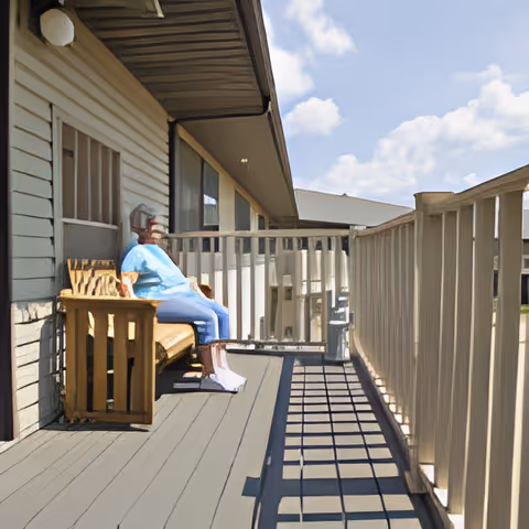 Sunlit second-story balcony with decking, railings, and a seated figure on a wooden bench beside the building.