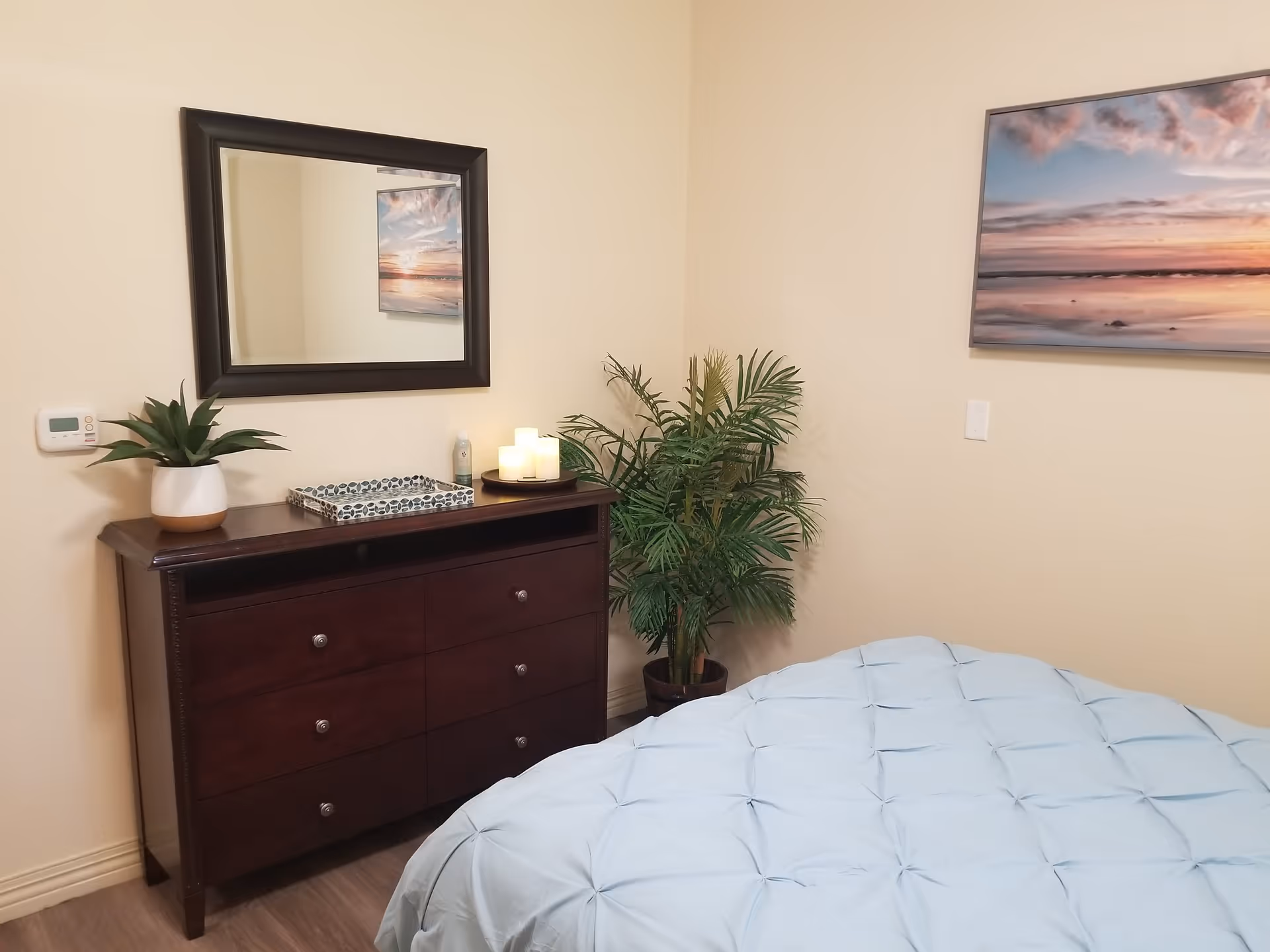 A bedroom corner with a dark wooden dresser topped with a potted plant, a decorative tray, and three lit candles. Above the dresser is a large rectangular mirror. To the right, there is a green potted plant and a wall-mounted picture of a sunset over the ocean. Part of a bed with a light blue comforter is visible in the foreground.