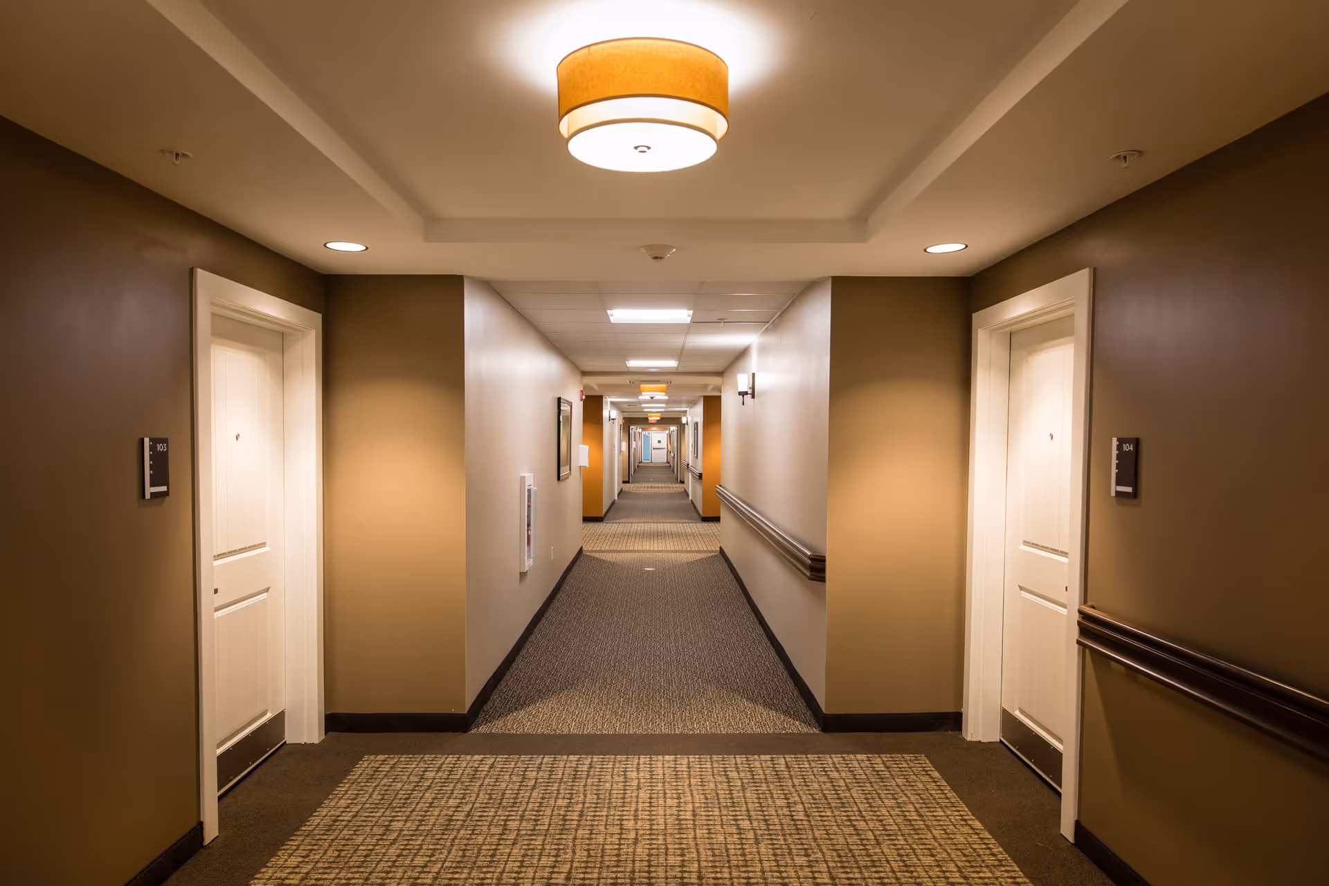 Well-lit carpeted interior hallway with numbered apartment doors, handrails, and ceiling lights.