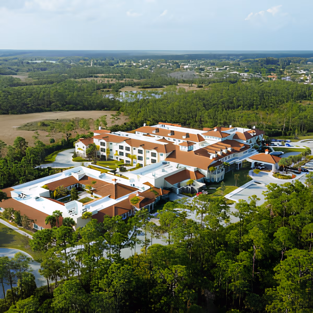 Aerial view of a large senior living facility with multiple connected buildings featuring white walls and reddish-brown roofs, surrounded by dense green trees and natural landscape under a partly cloudy sky.