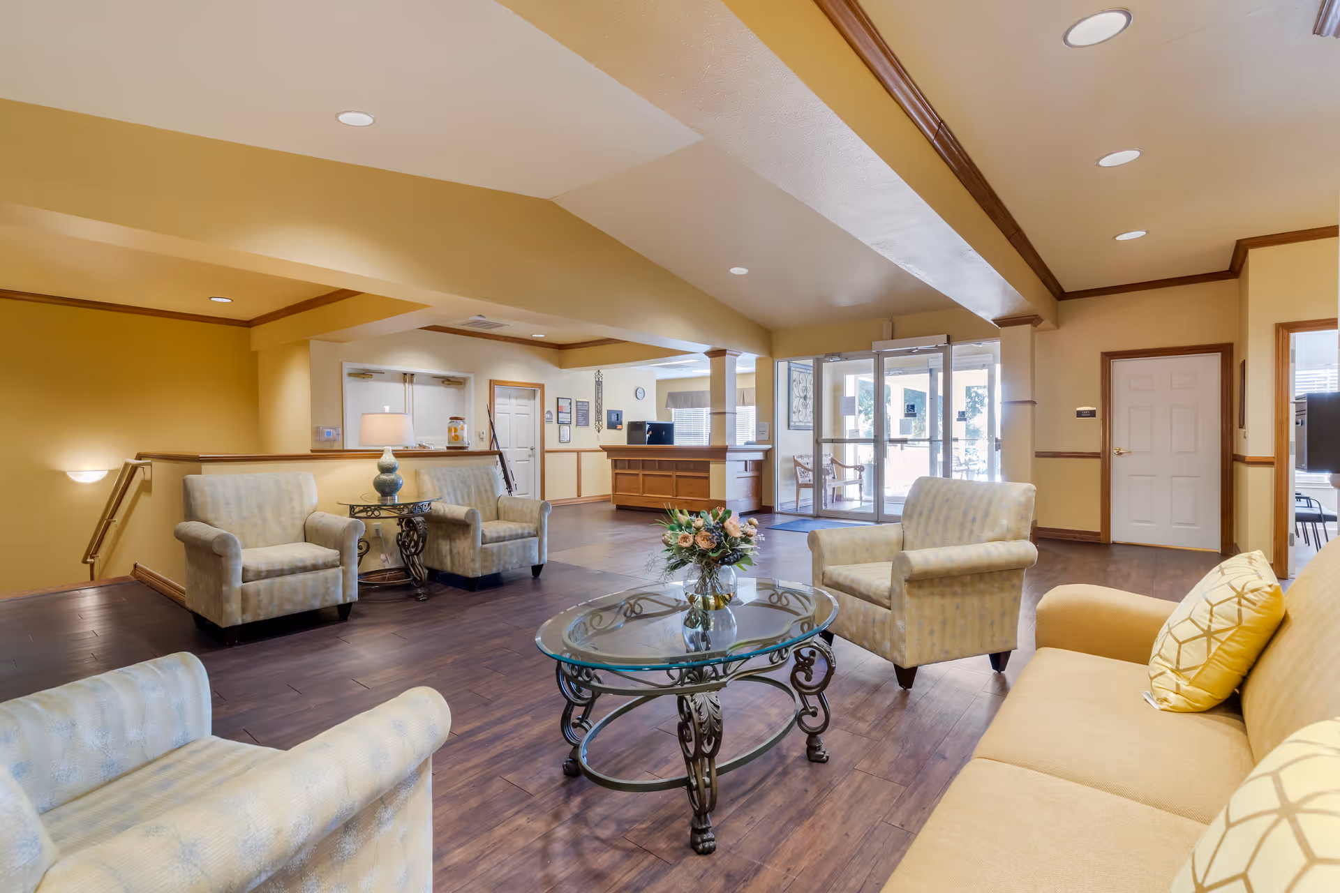 A bright and welcoming senior living facility lobby with beige walls and wooden flooring. The seating area includes a yellow couch with patterned pillows, three beige armchairs, and a glass coffee table with a floral arrangement. In the background, there is a reception desk and glass doors leading outside.
