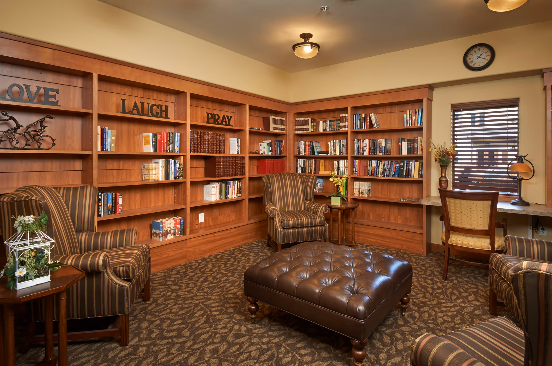 Cozy library room with wooden bookshelves filled with books and decorative words 'LOVE', 'LAUGH', and 'PRAY' on the top shelves. The room has striped armchairs, a large tufted leather ottoman in the center, a small side table with flowers, a desk with a chair and a lamp near a window with blinds, and a wall clock above the window.