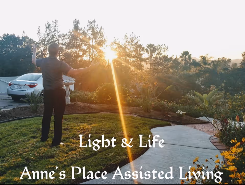 A person standing on a grassy area with arms outstretched facing the setting sun, surrounded by trees and plants, with a car parked on a driveway to the left. The scene is peaceful and bathed in warm sunlight.