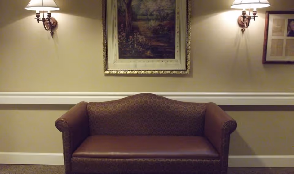 A brown upholstered loveseat with patterned fabric on the backrest and leather seat cushion is positioned against a beige wall. Above the loveseat hangs a framed painting depicting a nature scene. On either side of the painting are two wall-mounted lamps with white shades, casting warm light on the wall. There is a white chair rail molding running horizontally along the wall behind the loveseat.