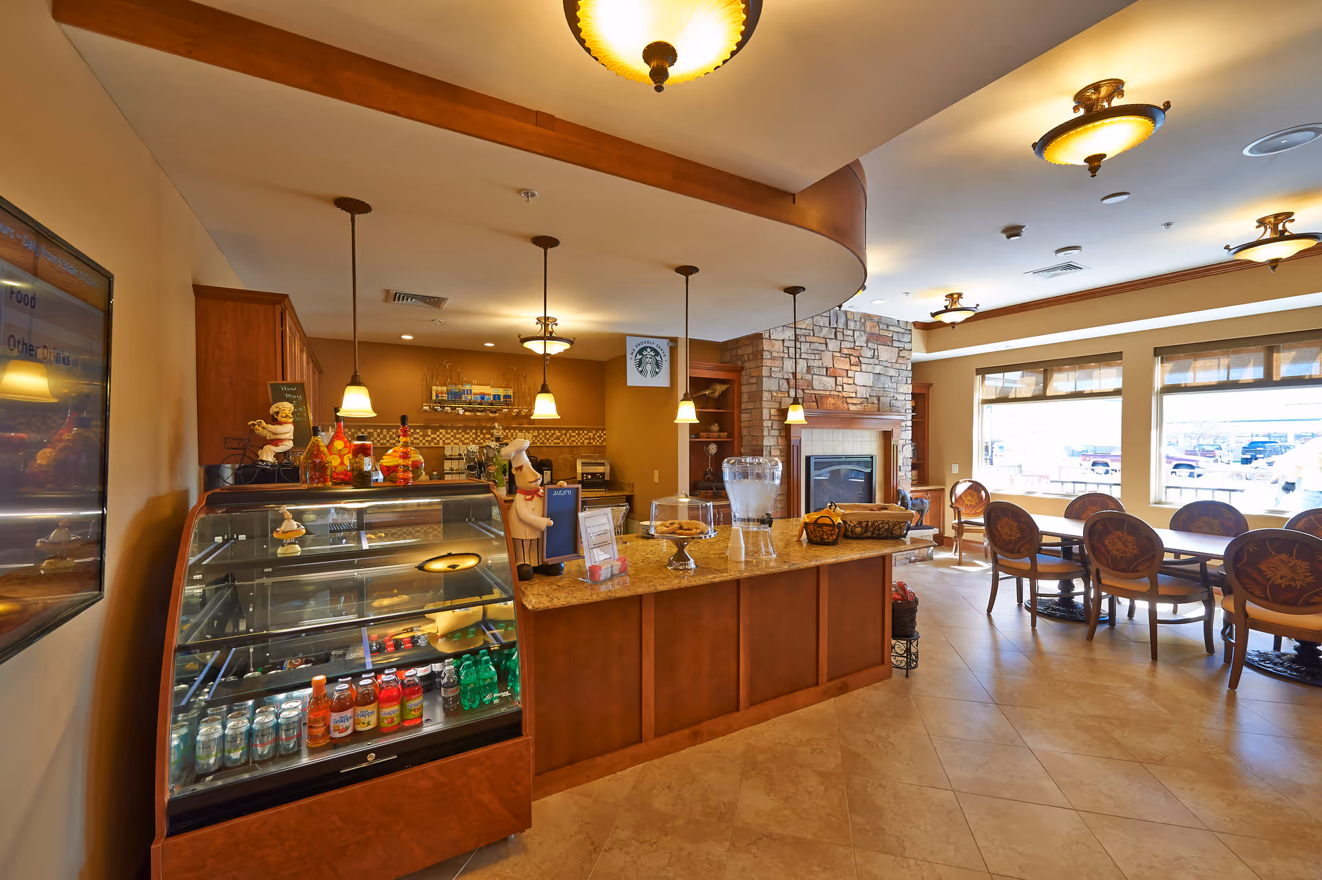 Interior view of a senior living facility cafe area with a glass display case containing beverages and snacks, a counter with decorative items and a water dispenser, pendant lights hanging from the ceiling, a stone fireplace in the background, and a seating area with tables and chairs near large windows letting in natural light.