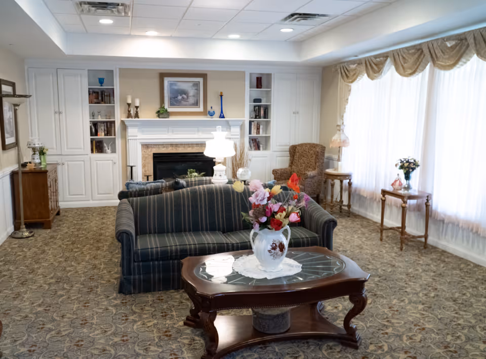 A cozy living room with a striped sofa facing a white fireplace with built-in shelves on either side. The room has patterned carpet, a wooden coffee table with a floral vase centerpiece, and large windows with sheer curtains and valances. There are also armchairs, side tables with lamps, and framed artwork above the fireplace.