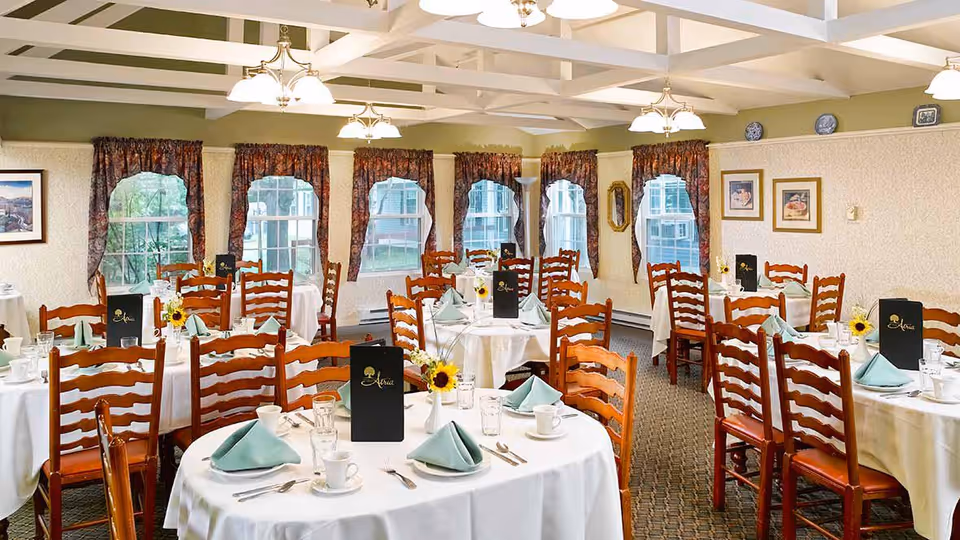 A bright dining room with multiple round tables covered in white tablecloths, each set with folded green napkins, cups, glasses, and silverware. Wooden chairs surround the tables, and menus with the name 'Atria' are placed on each table. The room has large windows with floral curtains, framed pictures on the walls, and ceiling lights with multiple bulbs providing warm illumination.