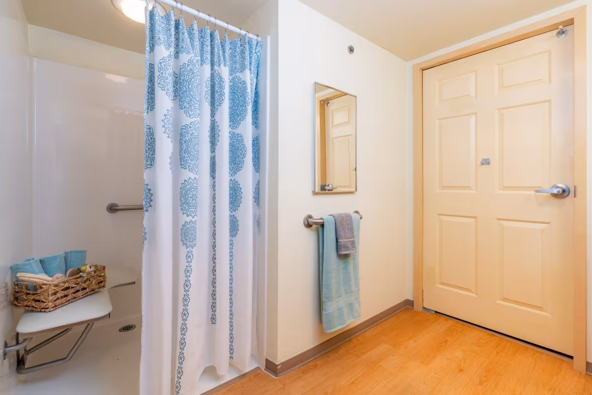 Small accessible bathroom featuring a shower with a blue-patterned curtain, fold-down seat and grab bar, a towel on a rail, and a closed door.