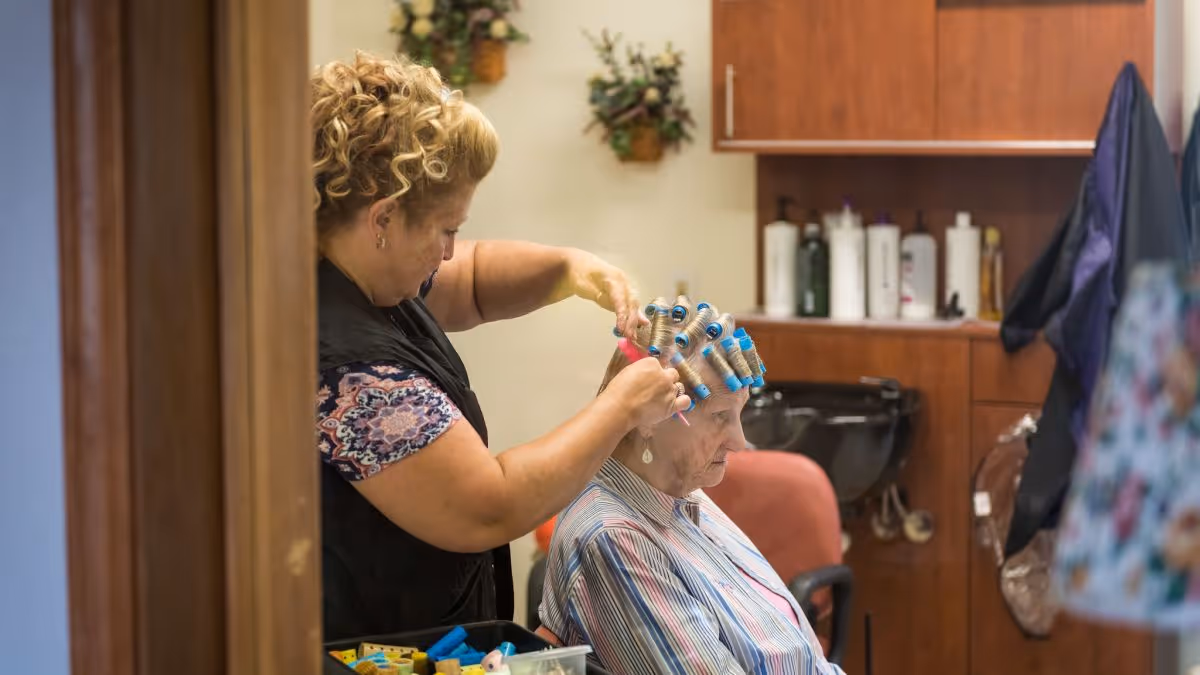 A woman is styling an elderly woman's hair with curlers in a salon-like setting. The elderly woman is seated wearing a striped shirt, while the stylist is standing behind her, focused on the hair. Shelves with hair care products and a hair washing station are visible in the background.