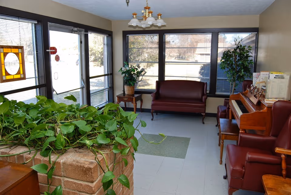 A bright room with large windows and blinds, featuring a maroon leather loveseat, matching armchairs, a wooden piano with sheet music, a small side table with a potted plant, and a brick planter filled with green leafy plants in the foreground.