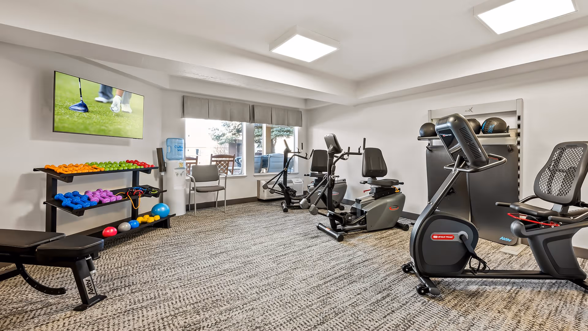 A bright exercise room with various fitness equipment including stationary bikes, a weight bench, colorful dumbbells on a rack, exercise balls, and a wall-mounted TV showing a golf game. There is a water cooler and chairs near a window with a view of outdoor greenery.