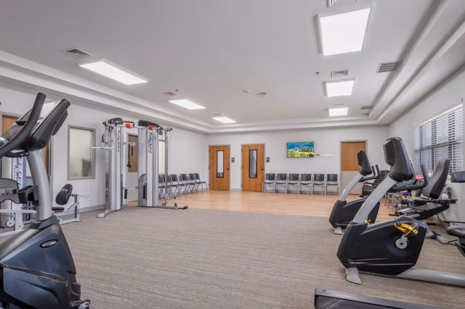 A spacious fitness room with exercise equipment including stationary bikes and a multi-functional weight machine. The room has a carpeted floor with a wooden section near the back, white walls, ceiling lights, and a row of chairs lined up against the far wall. There are three wooden doors and a window with blinds on the right side.