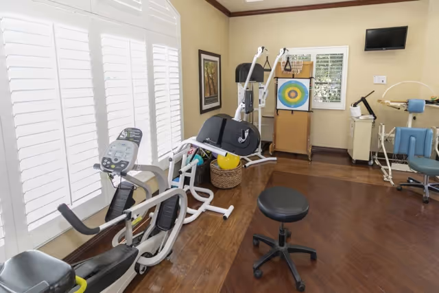 A bright rehabilitation room with stationary exercise equipment, therapy devices, a target board, and a rolling stool.
