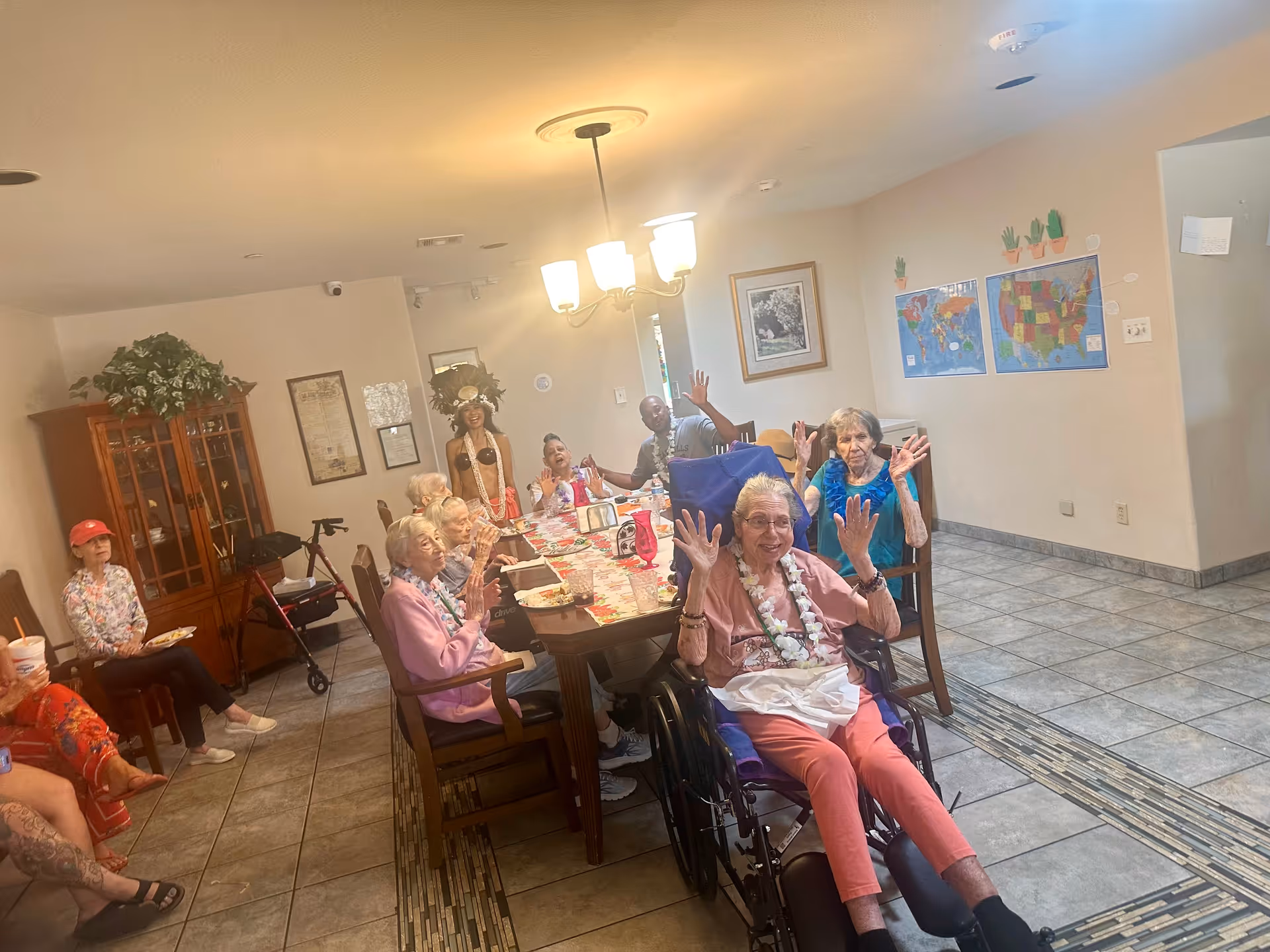 Seniors and staff gathered around a decorated communal dining table in a facility dining/activity room, several people waving at the camera.