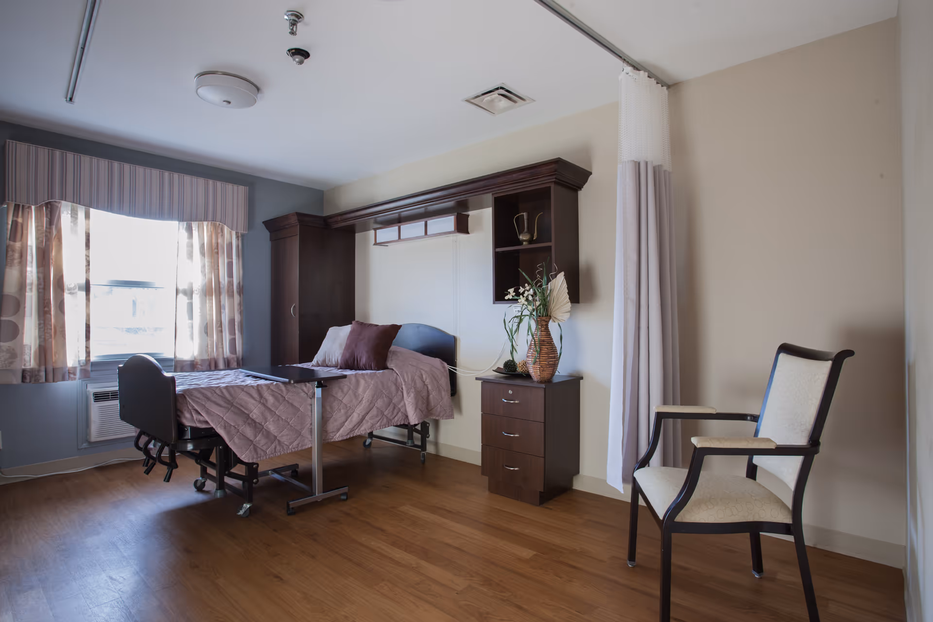 A furnished senior living bedroom with a hospital-style bed, nightstand, chair, and a window with curtains.