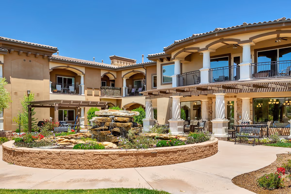 Outdoor courtyard area of The Retreat at Sunbrook featuring a stone water fountain surrounded by a circular raised flower bed with various plants and flowers. The courtyard is bordered by a two-story building with balconies, large windows, and a covered patio area with seating. The sky is clear and blue.