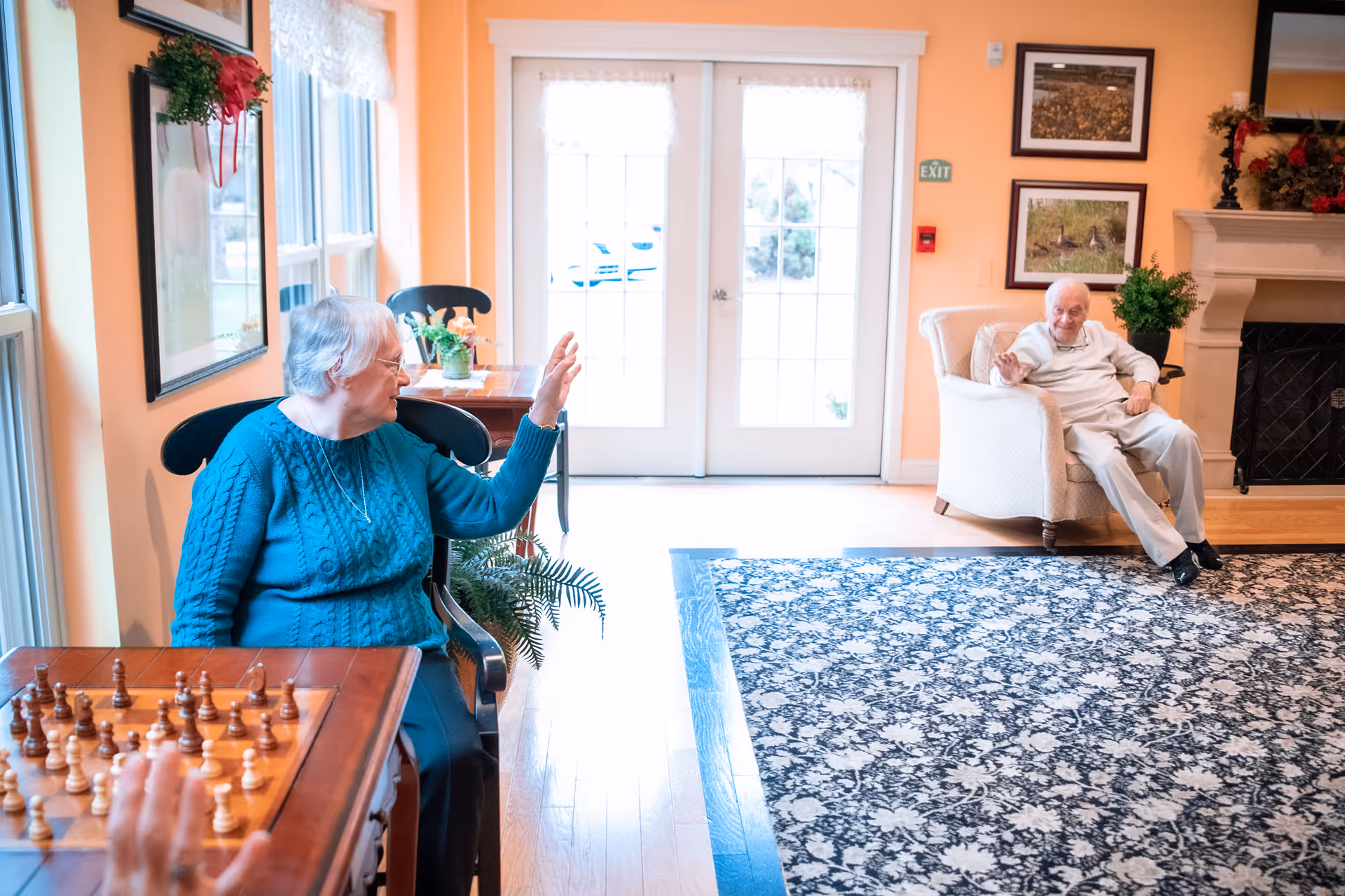 Two elderly individuals in a bright, cozy living room with peach-colored walls. One woman in a teal sweater is seated at a table with a chessboard, waving towards an elderly man sitting in a white armchair near a fireplace. The room has large windows, framed pictures on the wall, and a patterned rug on the wooden floor.
