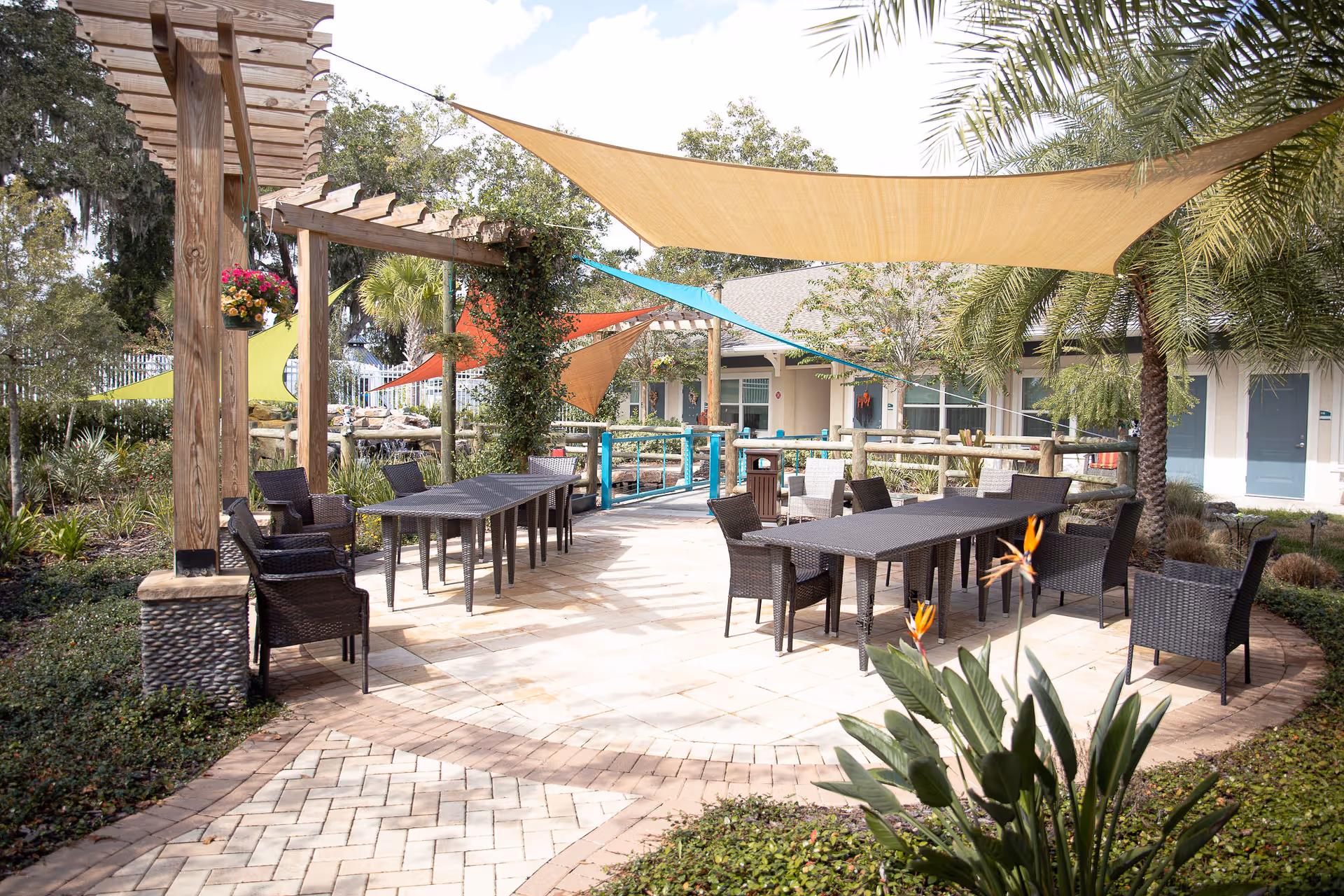 Outdoor patio with wicker tables and chairs under colorful shade sails and a pergola, surrounded by landscaping and a residential building.