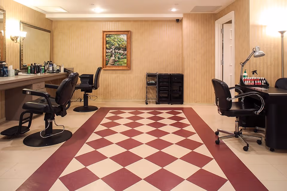 Interior view of a salon area with two black salon chairs on the left in front of a counter with hair products and a large mirror. On the right, there is a black chair at a manicure table with nail polish bottles and a desk lamp. The floor has a red and white diamond pattern, and a framed painting hangs on the beige wallpapered wall.