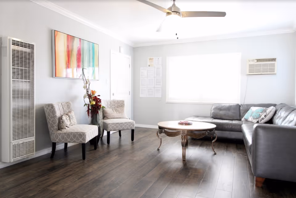 A bright living room with a gray leather sectional sofa adorned with decorative pillows, two patterned upholstered chairs with cushions, a round wooden coffee table, a tall vase with flowers, a wall-mounted heater, a colorful abstract painting, and a ceiling fan. The room has light gray walls and dark wood flooring.