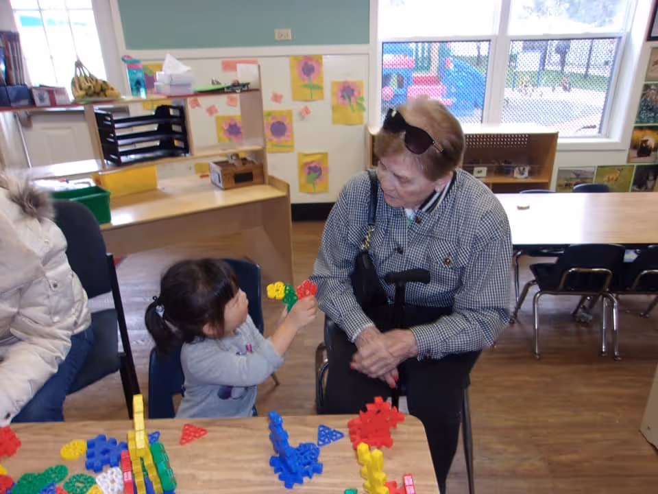 An elderly woman sitting at a table interacting with a young girl who is holding colorful plastic building pieces in a classroom or playroom setting. The room has children's artwork on the walls and large windows in the background.