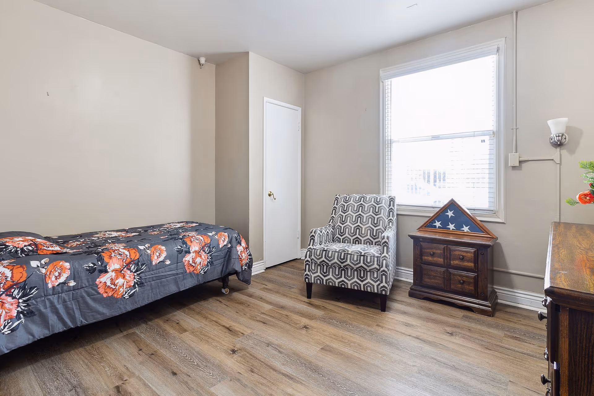 A simple bedroom with a single bed covered in a dark floral bedspread, a patterned armchair, a wooden cabinet with a folded American flag in a display case on top, a window with blinds, and a wooden dresser with a vase of flowers.