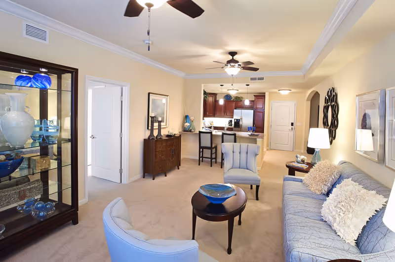 Open-plan living room featuring a sofa, armchairs, coffee table, display cabinet, and a view into the kitchen.