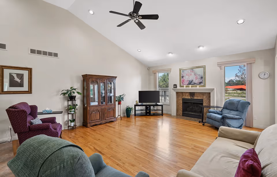 A spacious living room with wooden flooring, a ceiling fan, and recessed lighting. The room features a purple armchair, a blue recliner, a beige sofa with a purple pillow, a wooden cabinet with glass doors, a TV on a black stand, and a fireplace with a floral painting above it. Two windows with beige blinds allow natural light to enter, and there are some potted plants near the cabinet.