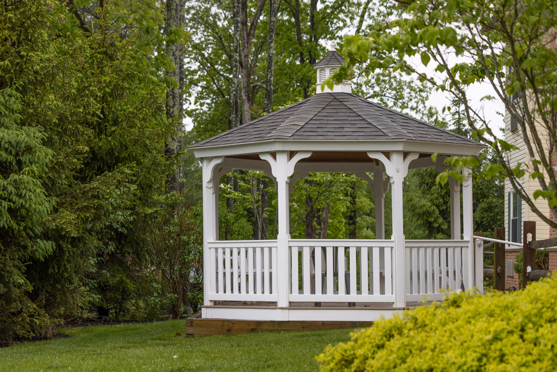 A white wooden gazebo with a shingled roof situated in a green garden area surrounded by trees and bushes, with part of a building visible on the right side.