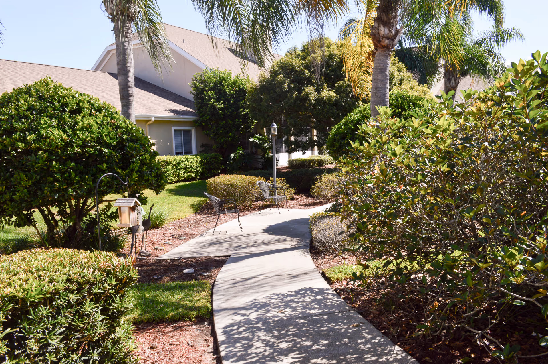 Curving concrete pathway through a landscaped garden with shrubs, palm trees, and a residential building in the background.