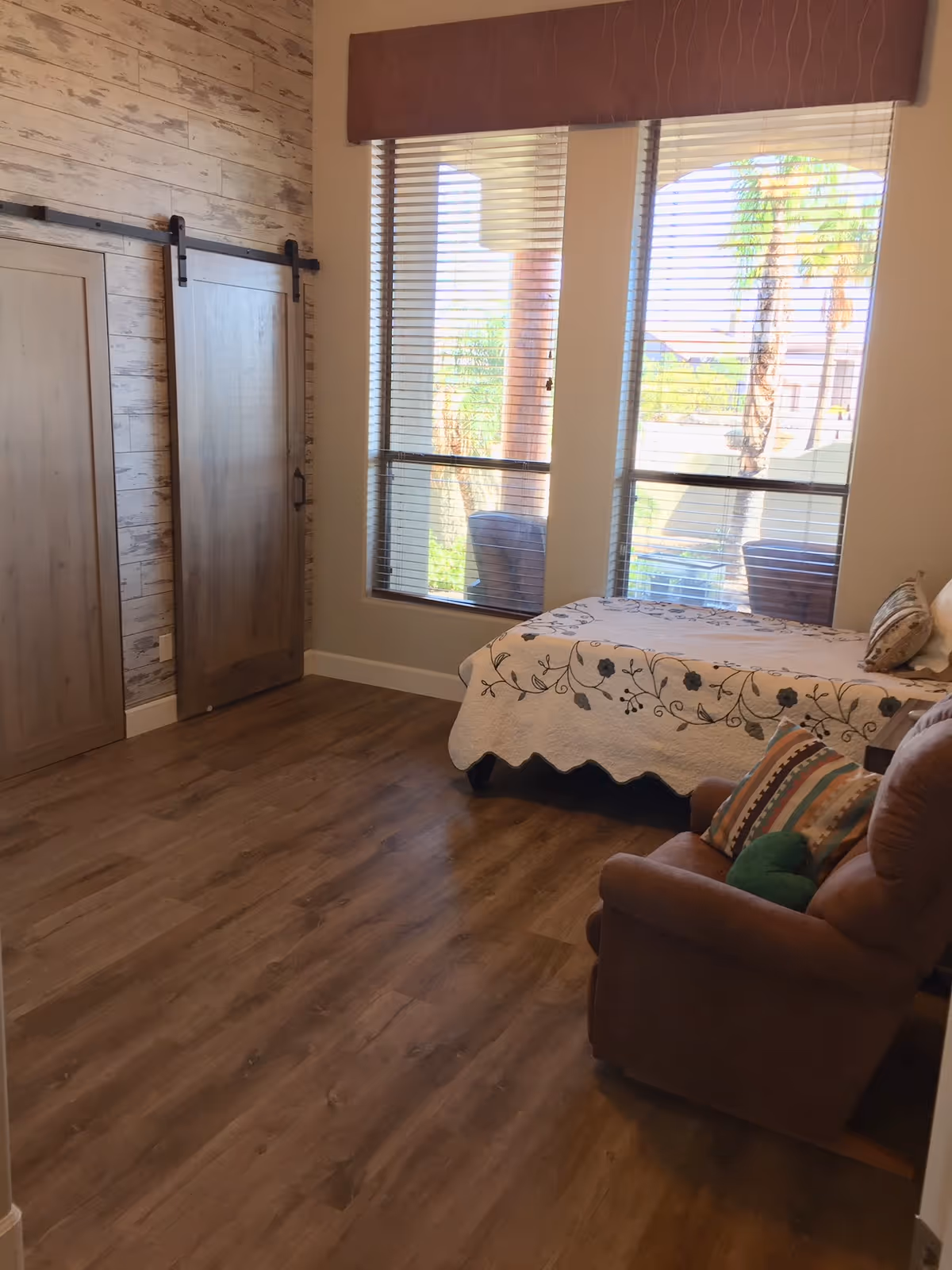 Bright bedroom with a single bed covered in a floral quilt, a recliner chair, wooden sliding barn doors, and large windows with blinds.