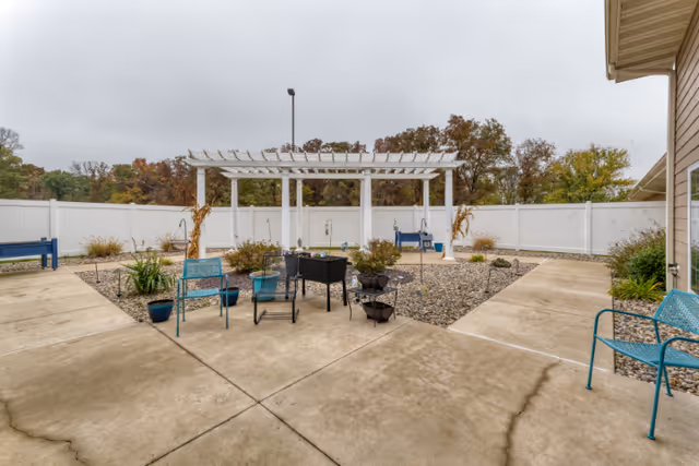 Outdoor patio area with concrete walkways, blue metal chairs, a white pergola, and various potted plants and shrubs. The area is enclosed by a white fence with trees visible in the background under an overcast sky.