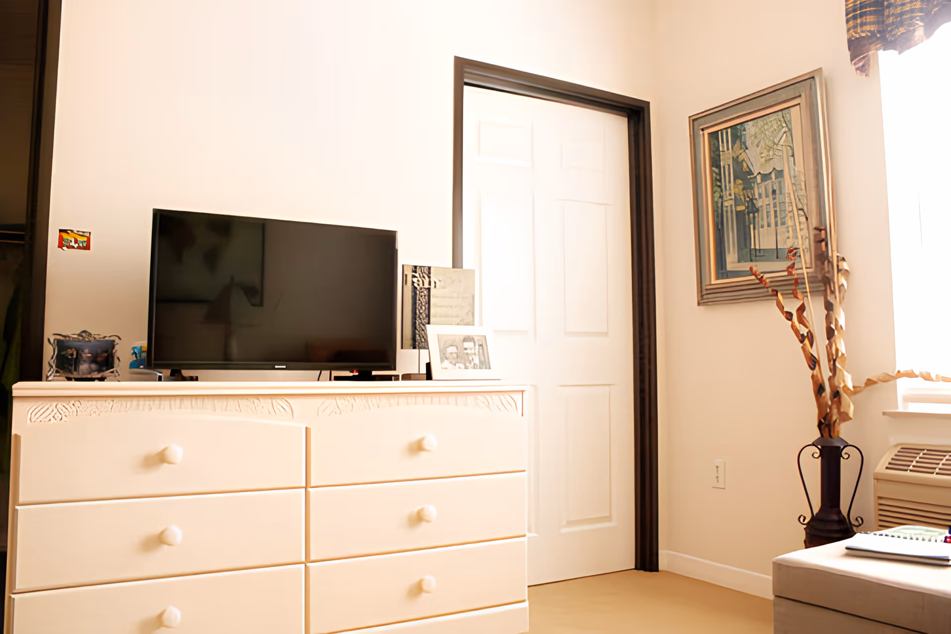 A bright room with a white dresser featuring six drawers and a flat-screen TV on top. Next to the TV are framed photos and a decorative item. A white door with dark trim is in the background, and a framed painting hangs on the wall to the right. A tall vase with dried decorative branches is placed near a window with a curtain and an air conditioning unit below it.