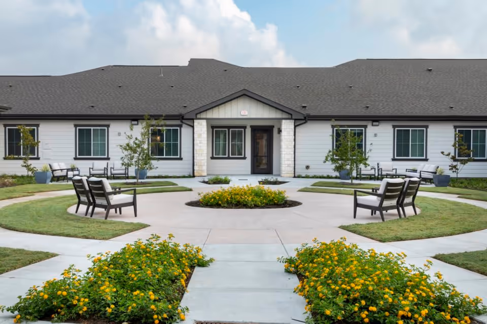 Outdoor courtyard area of a senior living facility with circular concrete pathways, green grass, yellow flowering plants, and several seating areas with cushioned chairs. The building in the background has white siding, black window frames, and a dark roof under a partly cloudy sky.