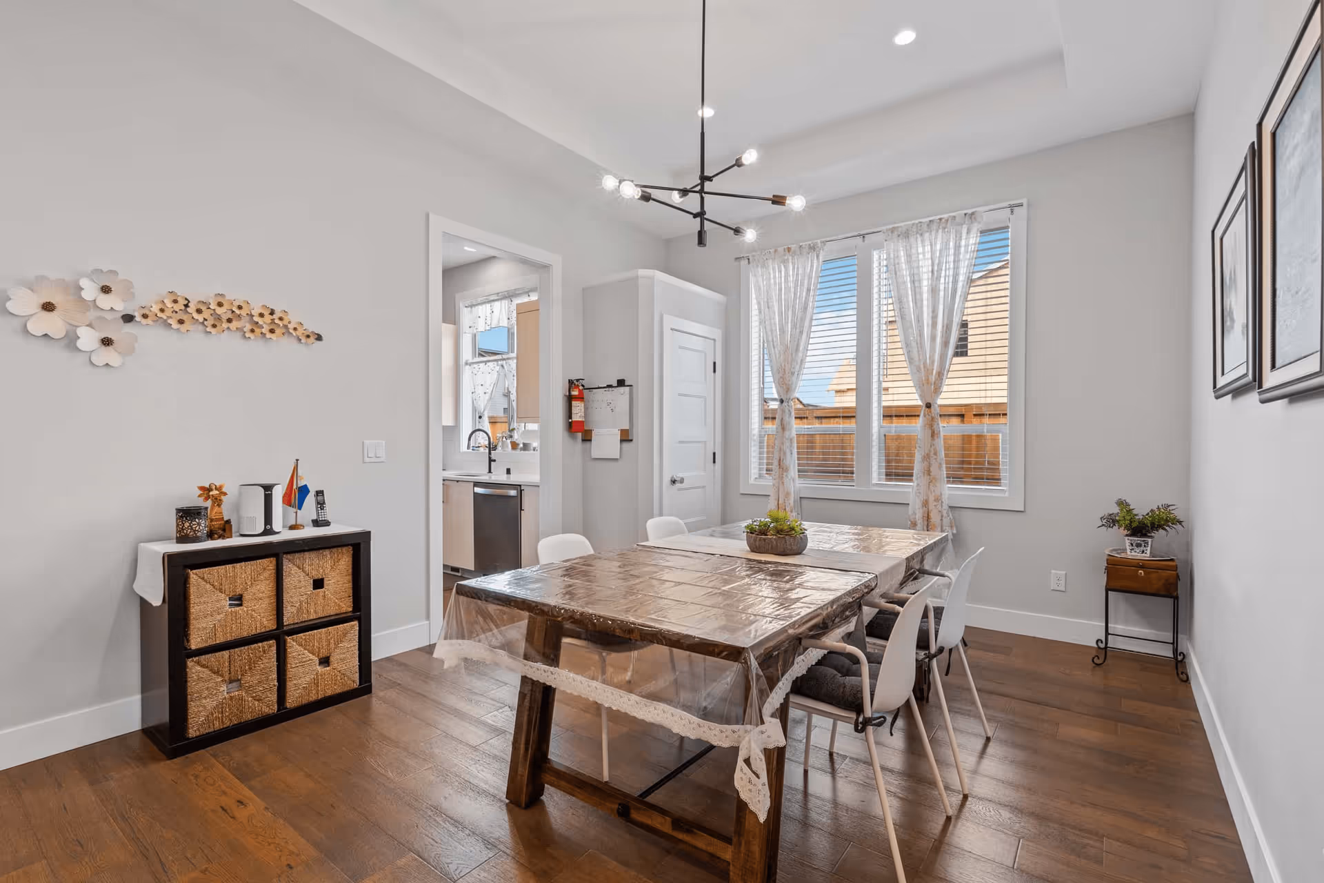 A bright dining room with a wooden table covered by a clear plastic tablecloth, surrounded by white chairs with cushions. The room has wooden flooring, a modern black chandelier with exposed bulbs, and large windows with sheer floral curtains letting in natural light. On the left wall, there is a small cabinet with woven baskets and decorative items on top, and a floral wall decoration above it. A doorway leads to a kitchen area with a window and stainless steel dishwasher visible. On the right wall, there are two framed pictures and a small table with a potted plant.