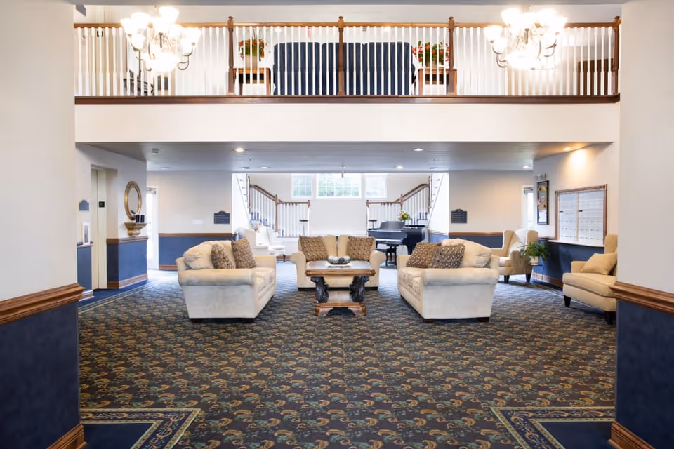 Spacious senior living facility common area with patterned carpet, three beige sofas arranged around a wooden coffee table, and additional armchairs along the walls. The room features a grand piano near the back, a staircase with white railings, and a balcony with seating and potted plants above. Two chandeliers hang from the ceiling providing warm lighting.