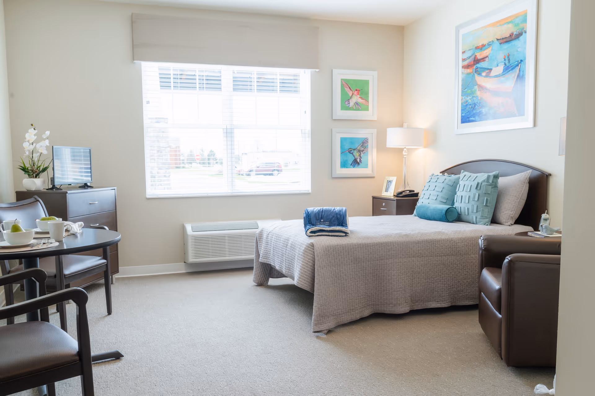 A bright and cozy bedroom in a senior living facility with a neatly made bed featuring light gray bedding and teal accent pillows. The room includes a brown leather armchair, a small round dining table with two chairs, a dresser with a small TV, and a window with white blinds letting in natural light. The walls are decorated with colorful framed artwork of birds and boats, and a table lamp sits on a nightstand beside the bed.