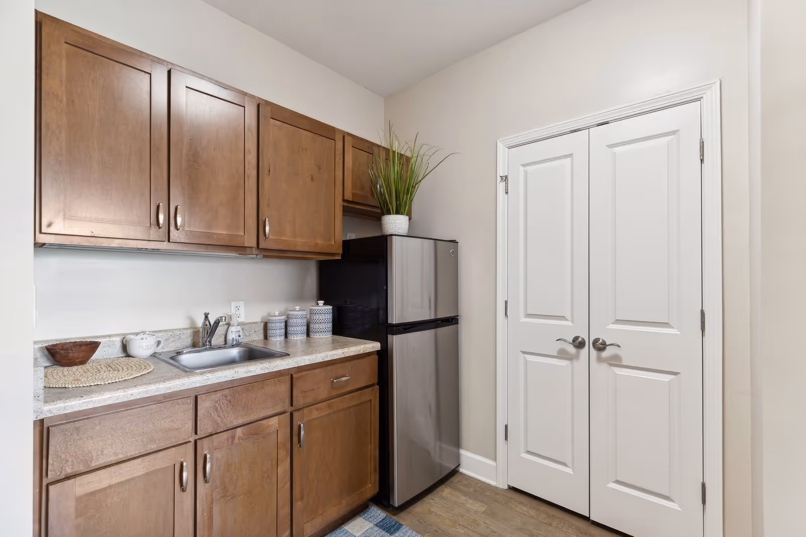 Small kitchen area with wooden cabinets, a stainless steel mini refrigerator, a countertop with a sink, and decorative jars. There is a potted plant on top of the refrigerator and a double white door on the right side.
