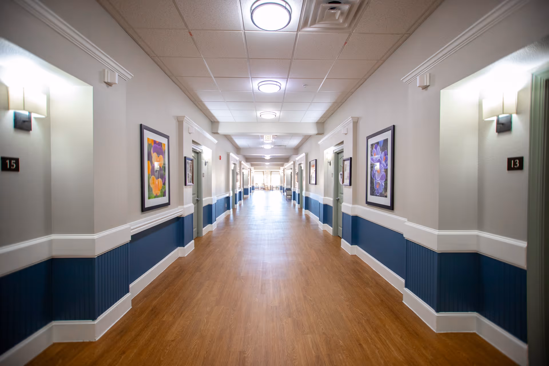 A long, well-lit hallway in a senior living facility with wooden flooring, blue and white walls, framed floral artwork, and numbered doors on each side.