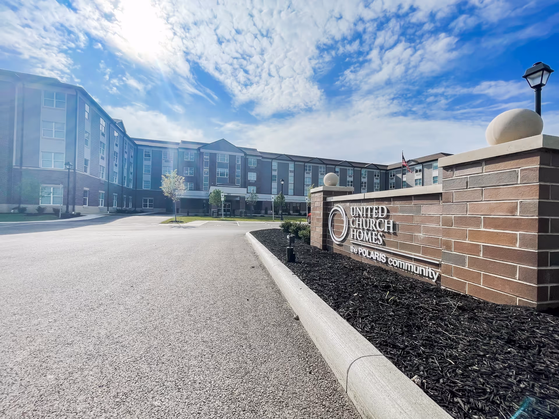 Exterior view of a multi-story senior living facility building under a partly cloudy sky, with a brick sign in the foreground that reads 'United Church Homes The Polaris Community'. The driveway and landscaped area with mulch and small trees are also visible.