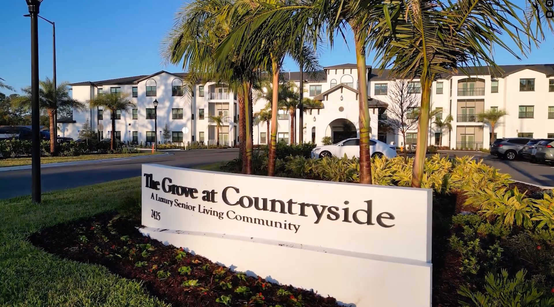 Exterior view of The Grove at Countryside, a luxury senior living community, showing a white multi-story building with balconies, palm trees, and a sign in the foreground with the community name and address.