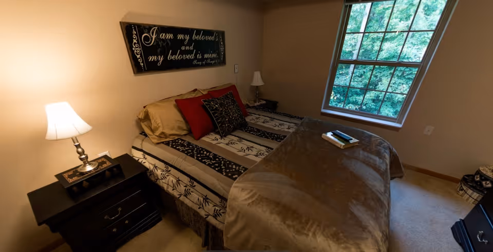 A cozy bedroom with a bed covered in a patterned bedspread and multiple pillows, two bedside tables each with a lamp, a large window showing green trees outside, and a decorative wall sign above the bed that reads 'I am my beloved's and my beloved is mine. Song of Songs 6:3'.