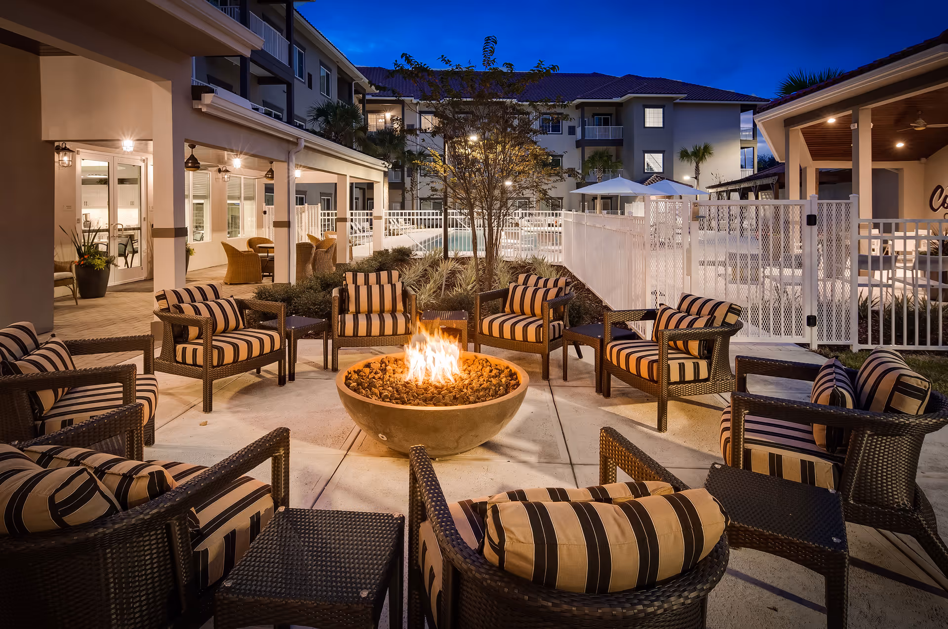 Outdoor courtyard at dusk with a central fire pit surrounded by striped cushioned chairs in front of a residential building.