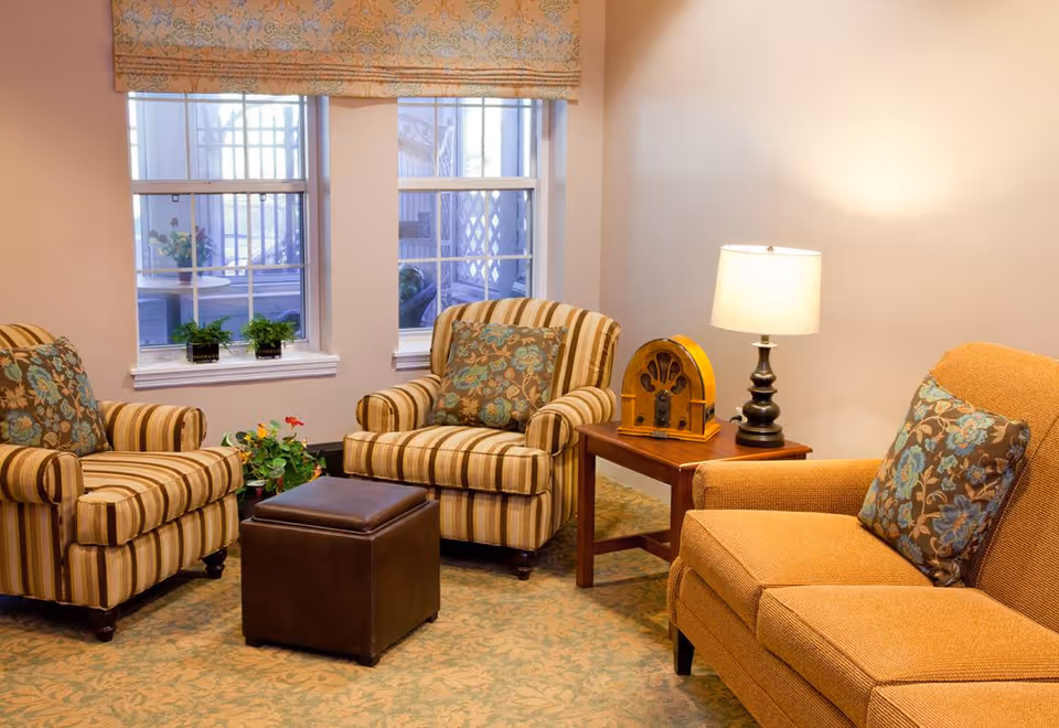 Cozy seating area with two striped armchairs, an upholstered sofa chair, a leather ottoman, a side table with a lamp and decorative radio, and windows behind.
