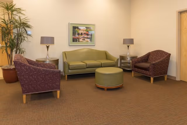A seating area with a green sofa, two patterned armchairs, and a round green ottoman in the center. There are two side tables with lamps on either side of the sofa, a potted plant in the corner, and a framed painting on the wall behind the sofa.
