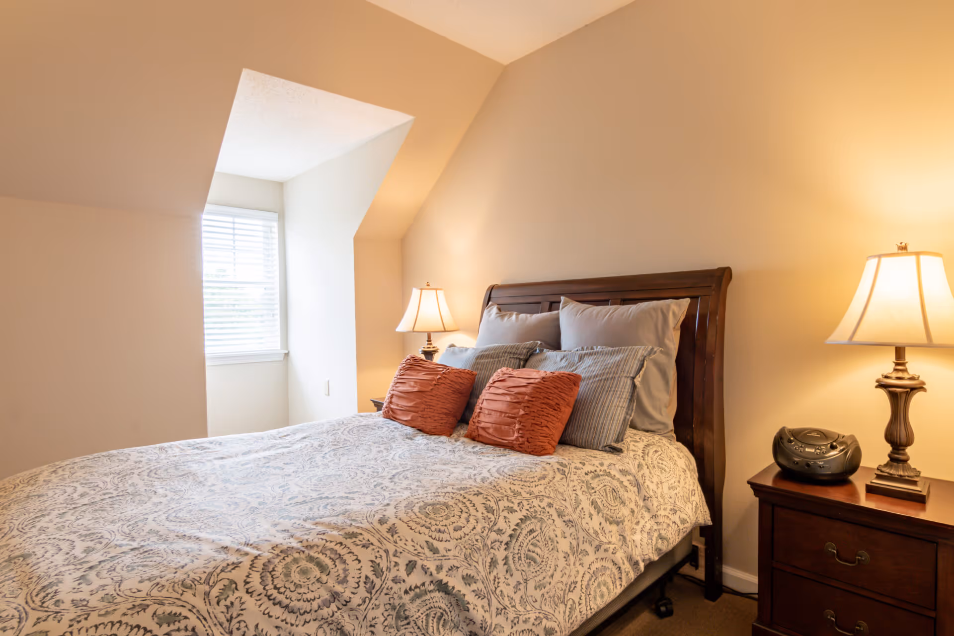 A cozy bedroom with a wooden bed frame, patterned bedspread, and multiple pillows including two orange decorative pillows. There are two bedside tables each with a lamp, and a small radio on the right bedside table. A window with white blinds is visible on the left side of the room.