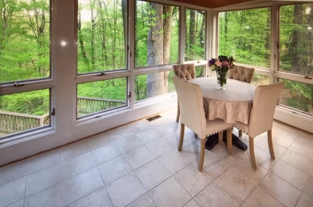 A bright dining area with large windows on three sides offering a view of lush green trees outside. The room features a round table covered with a beige tablecloth, surrounded by four beige upholstered chairs. A vase with flowers is placed in the center of the table.